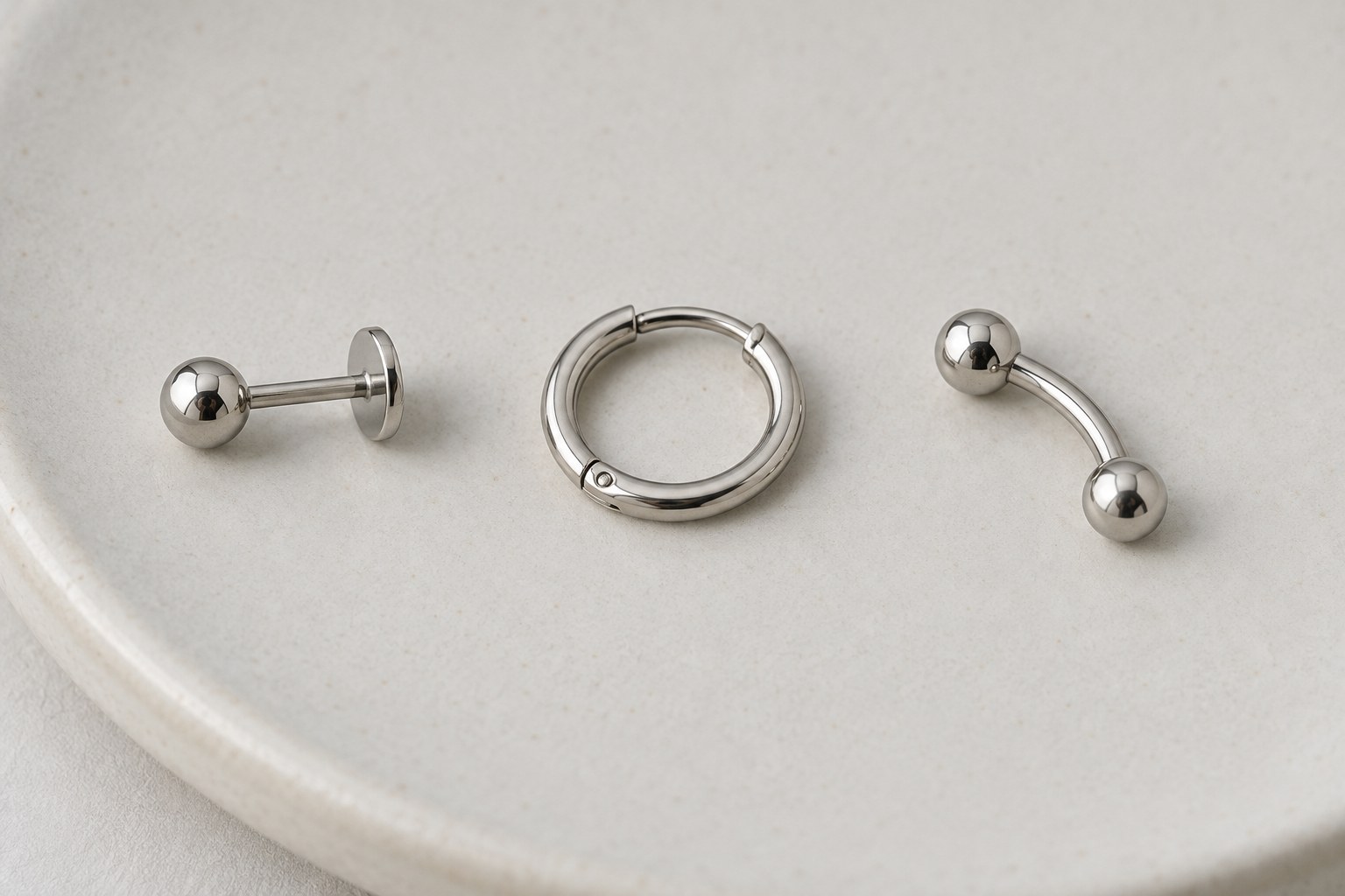 A flat-back stud, small hoop and curved barbell arranged separately on pale ceramic.