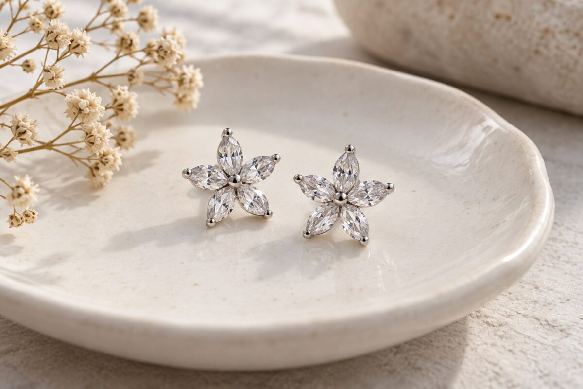 Two five-petal crystal flower studs on a pale dish beside a dried botanical sprig.