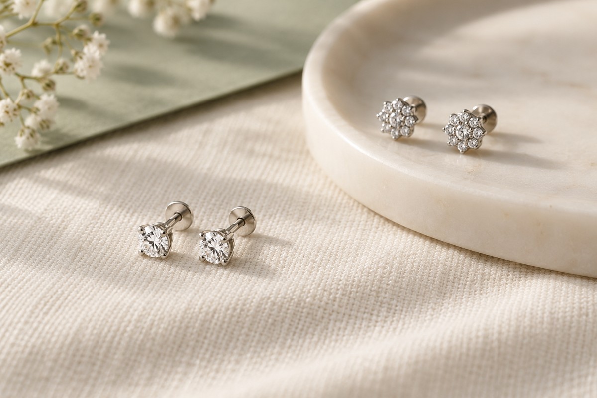 Two pairs of small clear-stone studs beside an ivory dish on cream fabric.